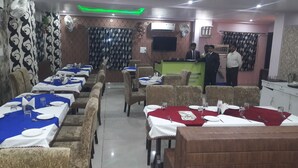 A restaurant with multiple tables, some with blue and red tablecloths, and a counter area with a person standing. - M S Hotel (Bhagalpur)