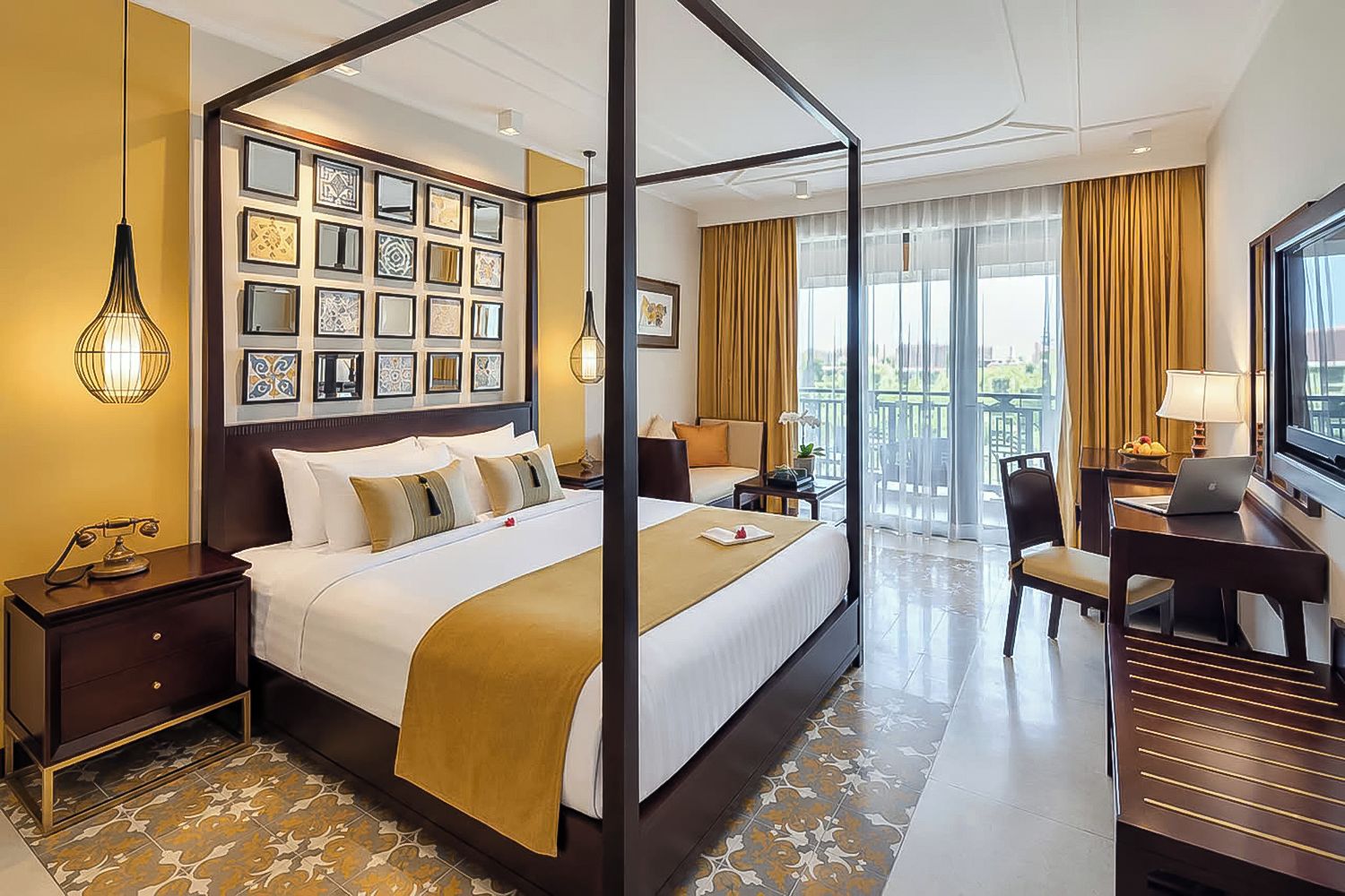 A hotel room with a four-poster bed, a desk with a laptop, a television, and a balcony with a view.