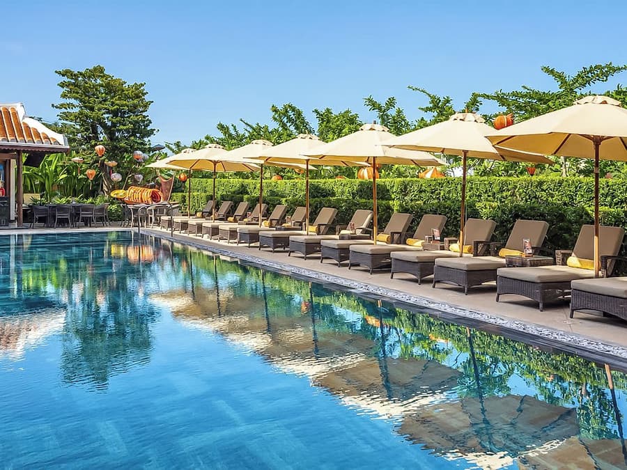 Una piscina con sillones de descanso y sombrillas, rodeada de follaje.