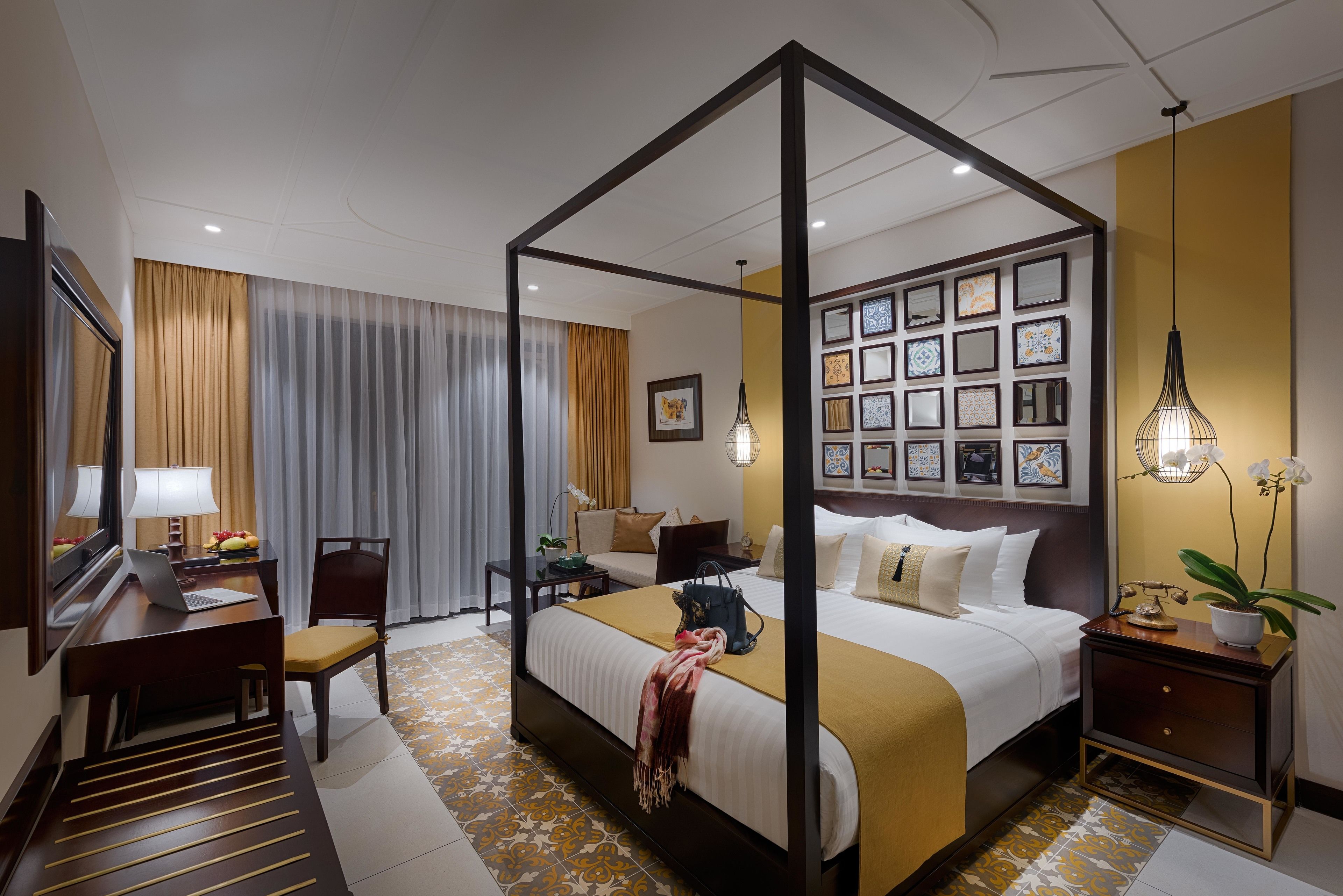 A four-poster bed with a headboard, a desk with a laptop, a chair, a bedside table with a lamp, and a wall with framed pictures.