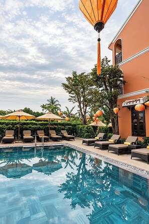 Ein Poolbereich mit Liegestühlen und Sonnenschirmen, umgeben von Grünflächen und einem Gebäude mit Balkon.