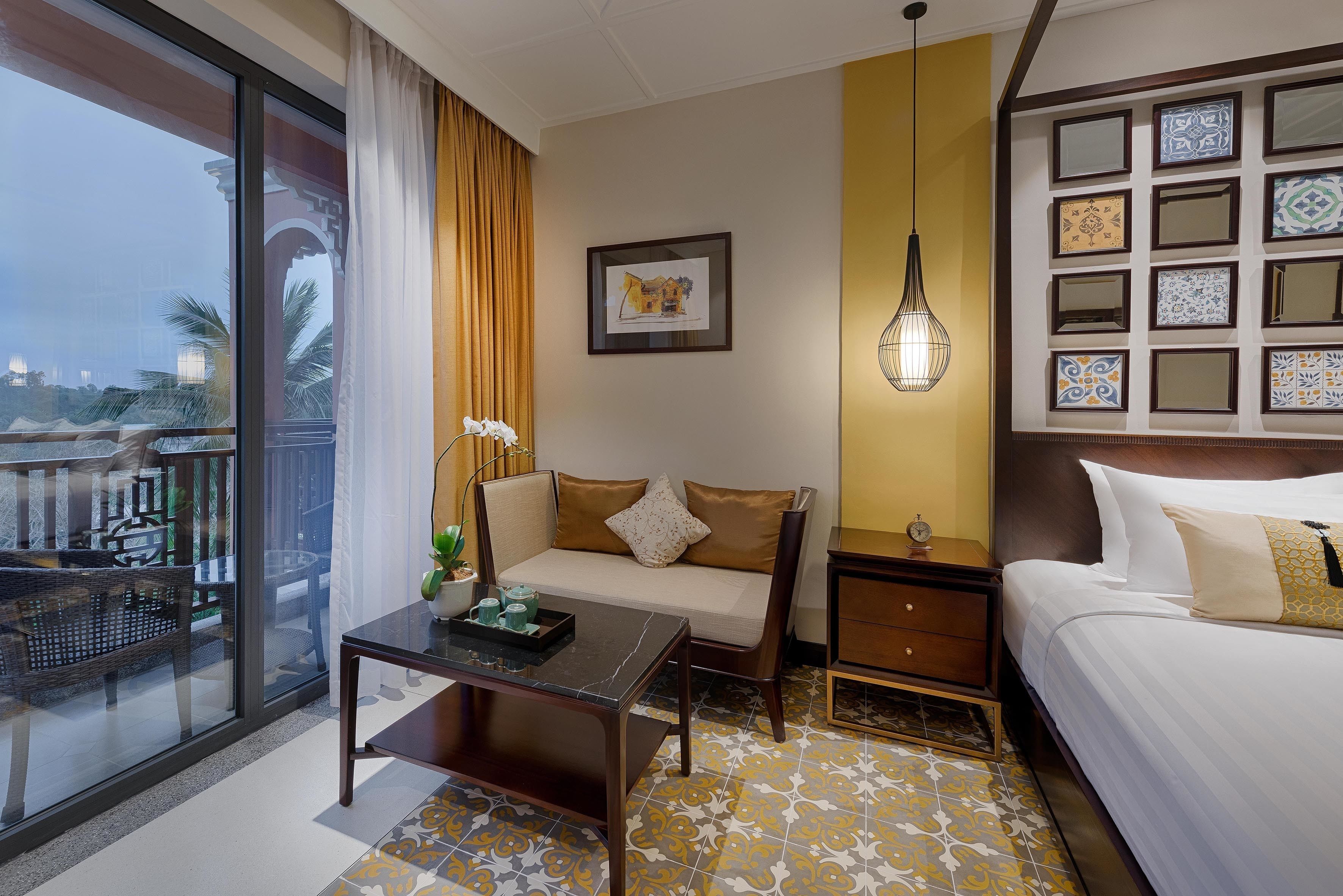 A hotel room with a bed, a sofa, a nightstand, a table with a tray, a potted plant, a framed picture, and a view of the outside.