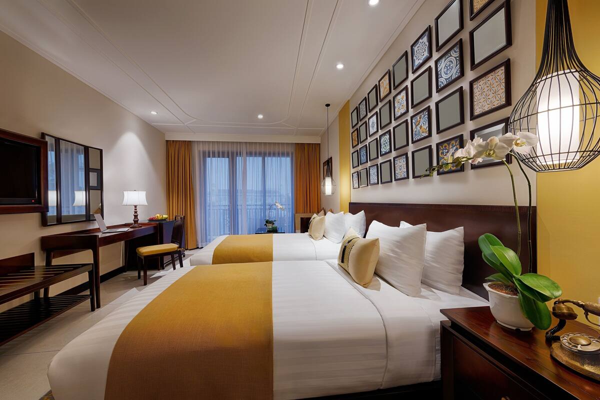 Habitación de hotel con dos camas, un escritorio con una computadora portátil, una silla, una lámpara, una planta en maceta y un cuadro con fotografías.