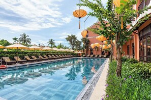 Ein Swimmingpool mit Liegestühlen und Sonnenschirmen, umgeben von viel Grün und einem Gebäude mit großen Fenstern.
