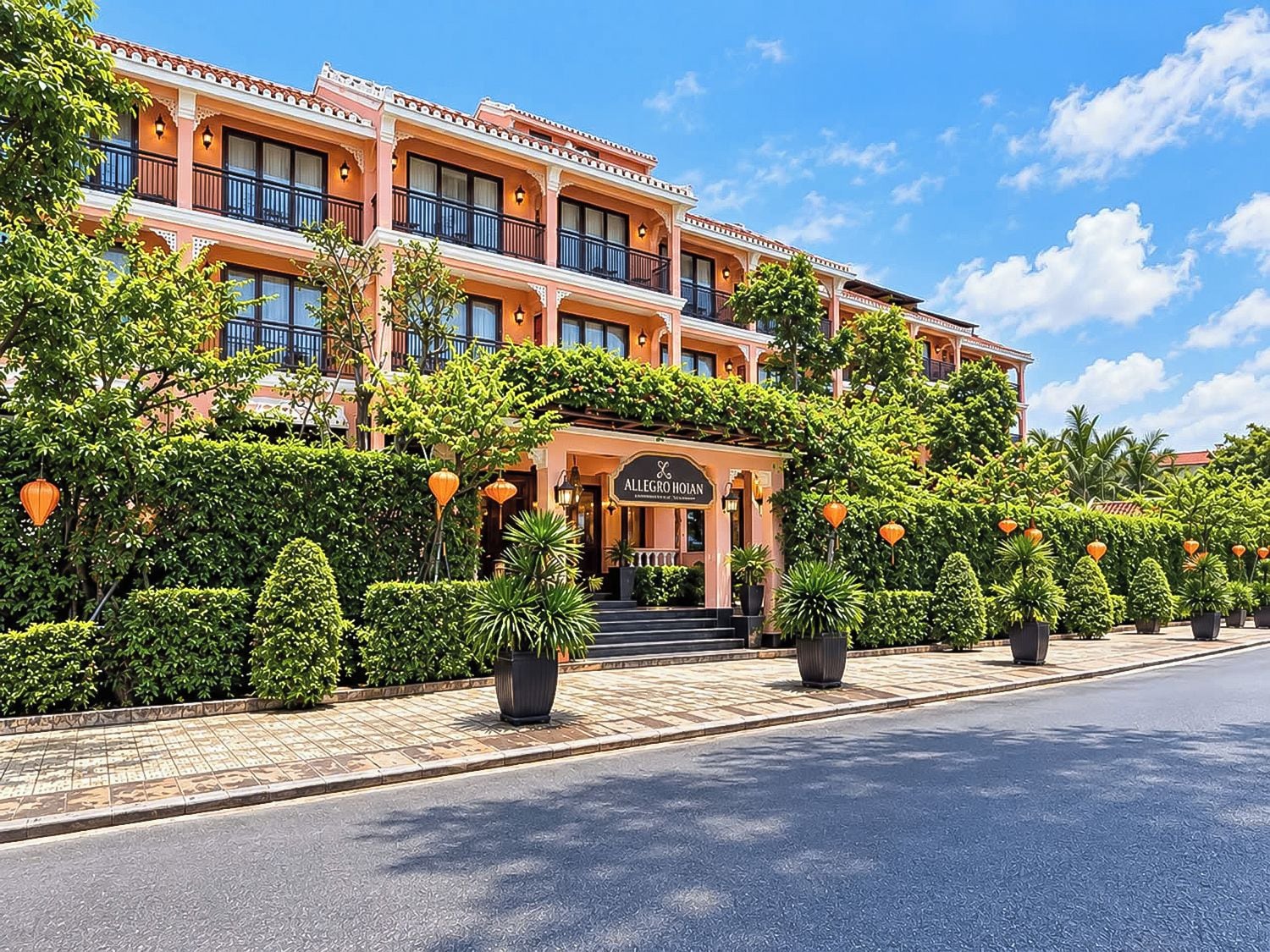 A multi-story hotel with a traditional architectural style, featuring a prominent entrance with a sign, and a well-maintained garden with potted plants and a paved walkway.