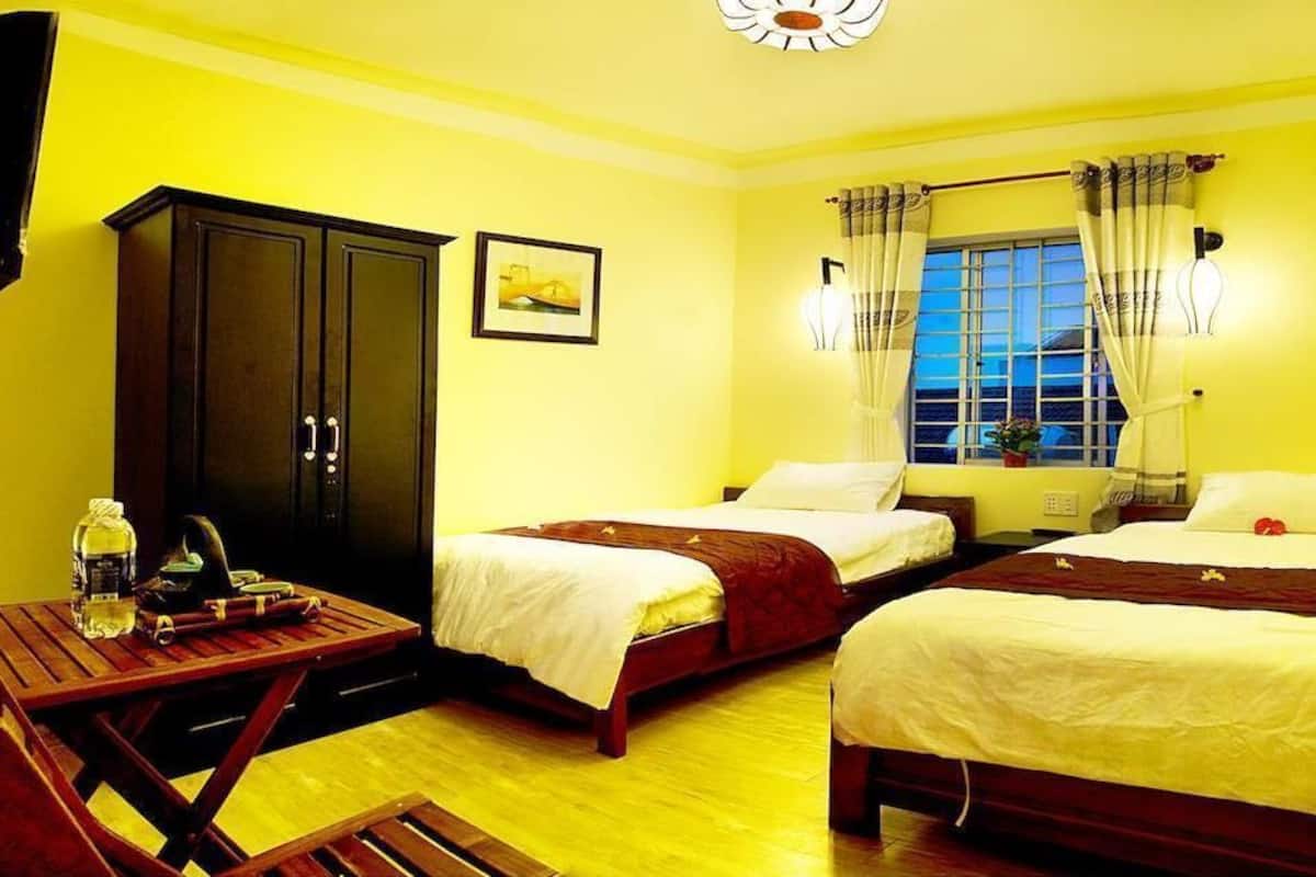 A hotel room with two single beds, a wooden table with a chair, a wardrobe, and a window with curtains.