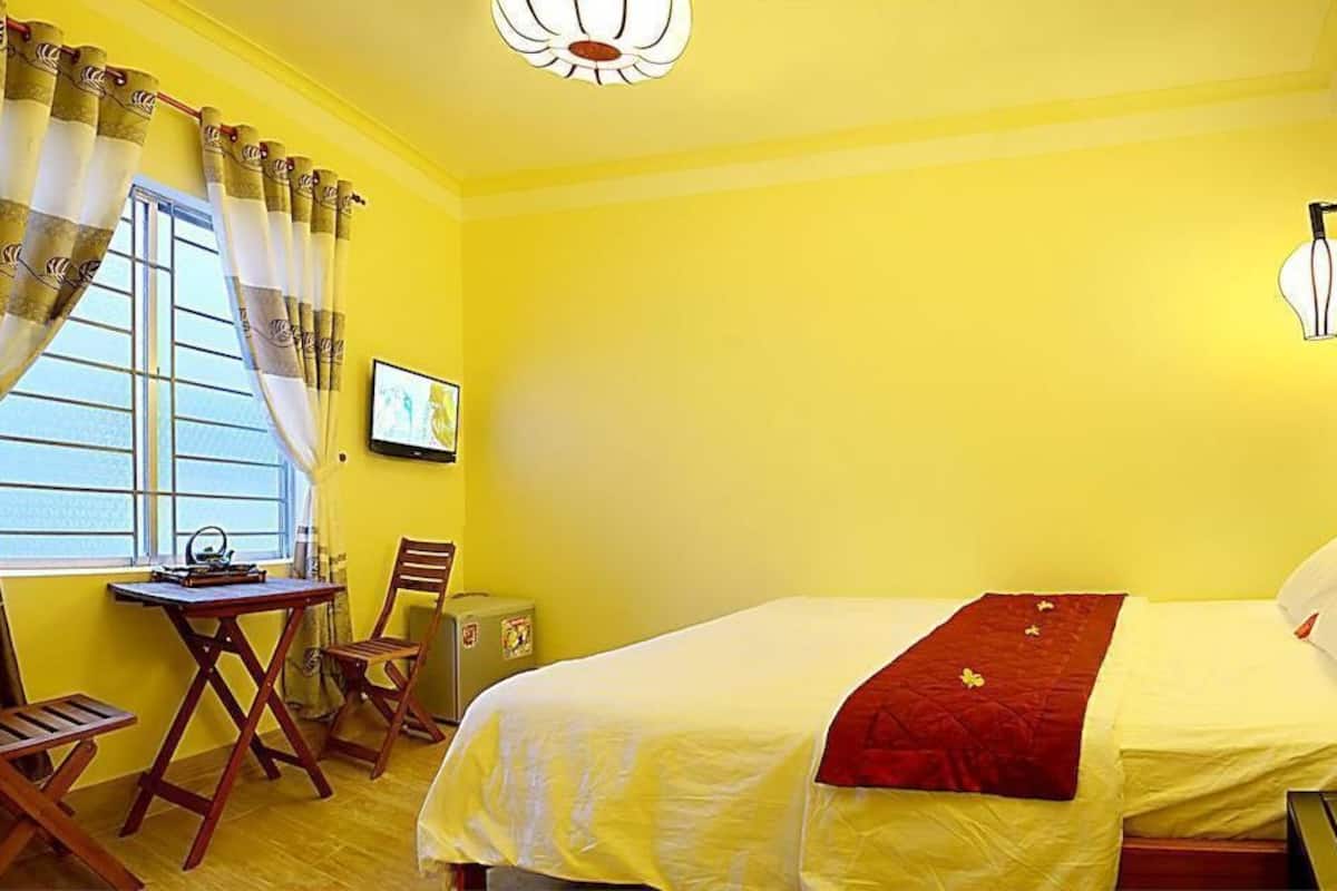 A room with yellow walls, a bed with a red blanket, a desk with a chair, and a window with curtains.
