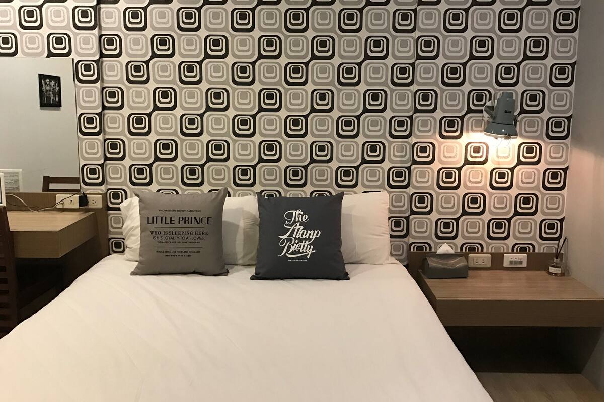 A bedroom with a patterned wallpaper, two bedside tables, a lamp, and two pillows with text.