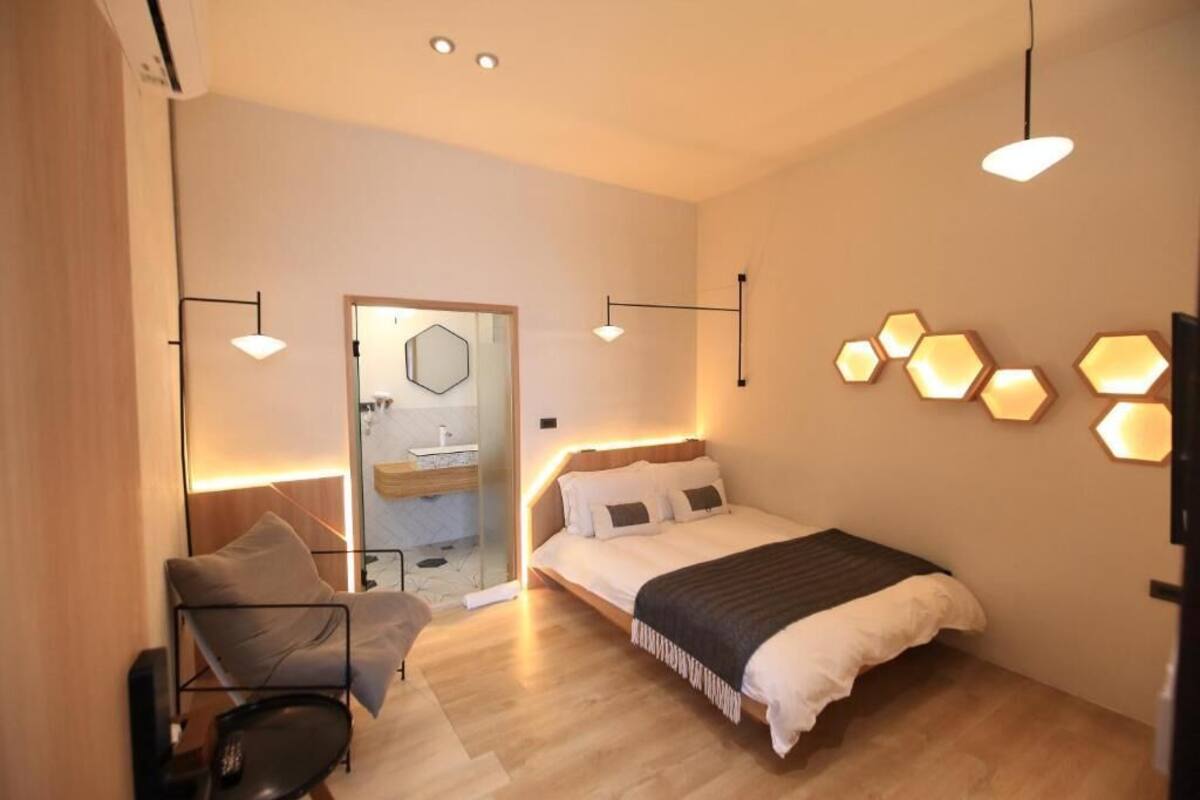 A modern bedroom with a bed, a chair, and a desk. There is a bathroom visible through a glass door.