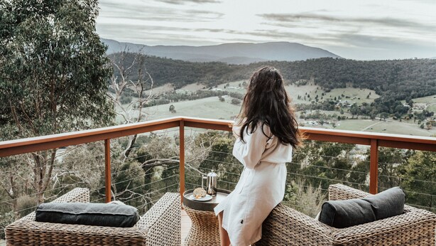 A woman in a white robe sitting on a wicker chair overlooking a scenic landscape with mountains and trees. - Kangaroo Ridge Retreat (Healesville)