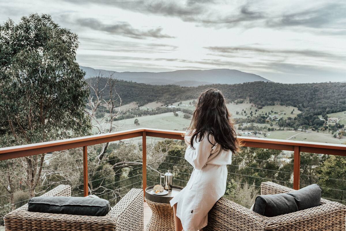 A woman in a white robe sitting on a wicker chair overlooking a scenic landscape with mountains and trees.