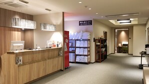 Uma área de recepção de hotel com um balcão de madeira, um computador e um refrigerador Coca-Cola. Há prateleiras com toalhas dobradas e uma placa em japonês.