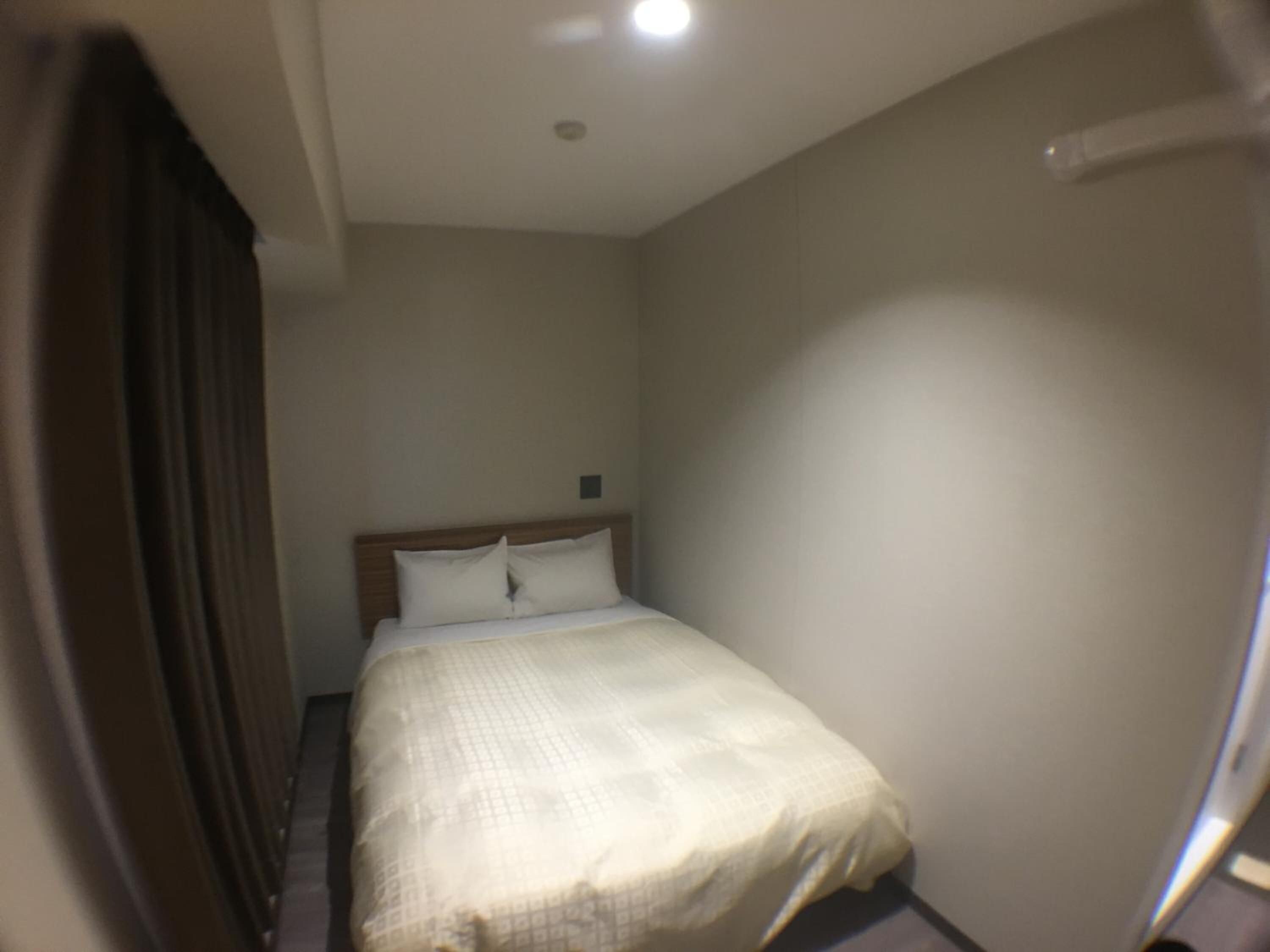 A narrow hotel room with a single bed, a small bedside table, and a plain wall.