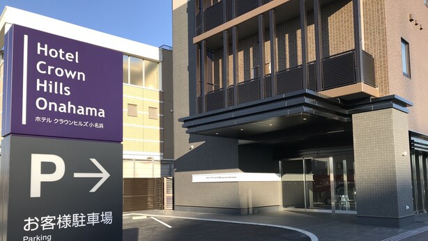 Entrada de hotel com placa indicando Hotel Crown Hills Onahama, sinal de estacionamento e um edifício com design moderno.