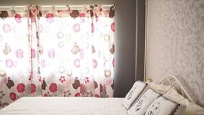 A neatly made bed with a quilted white cover, two pillows with a floral pattern, and a window with floral curtains. - Baan Tukta at Beach (Cha-am)