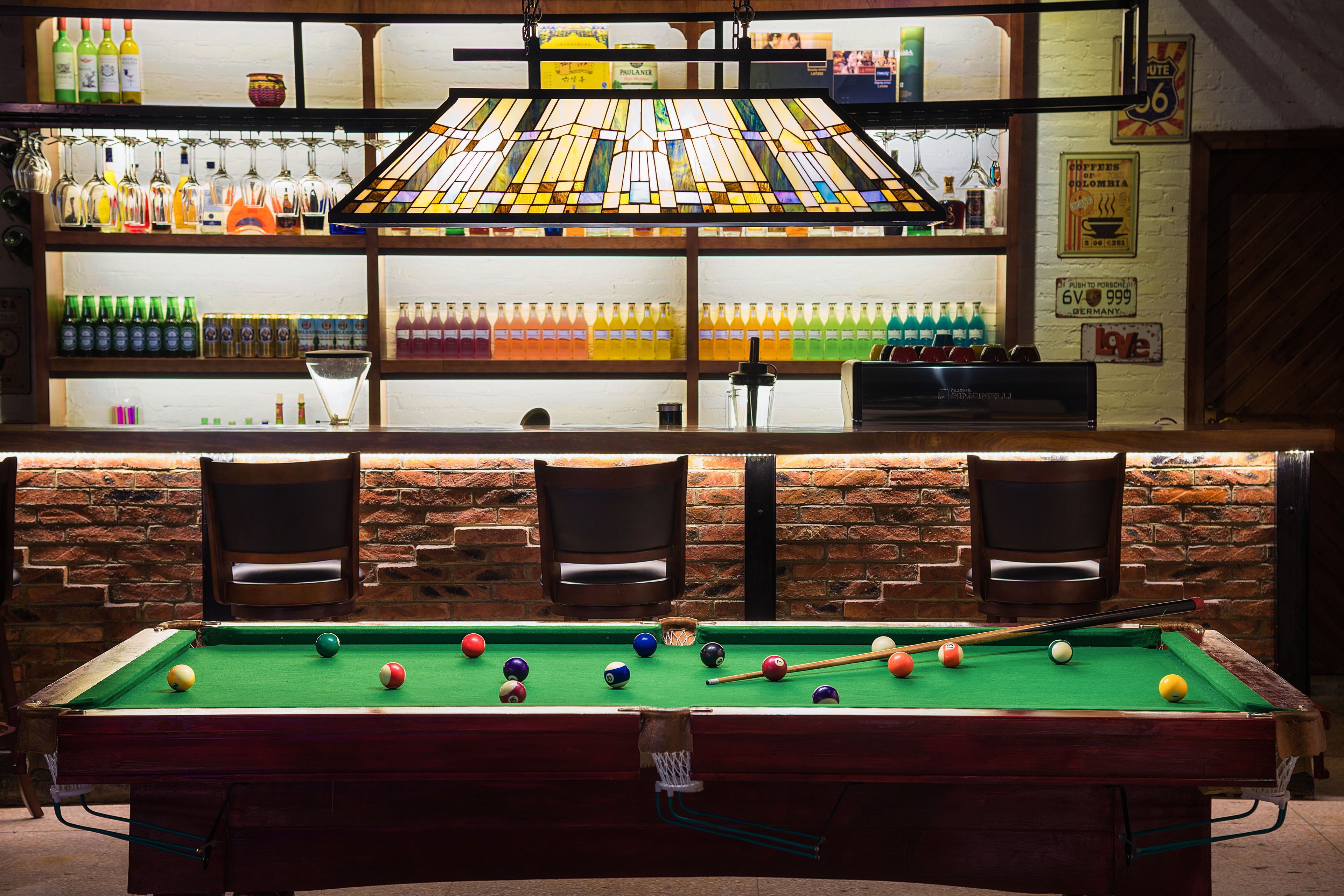 Une table de billard avec des billes et des queues, un bar avec des bouteilles et des verres, et des murs en briques.