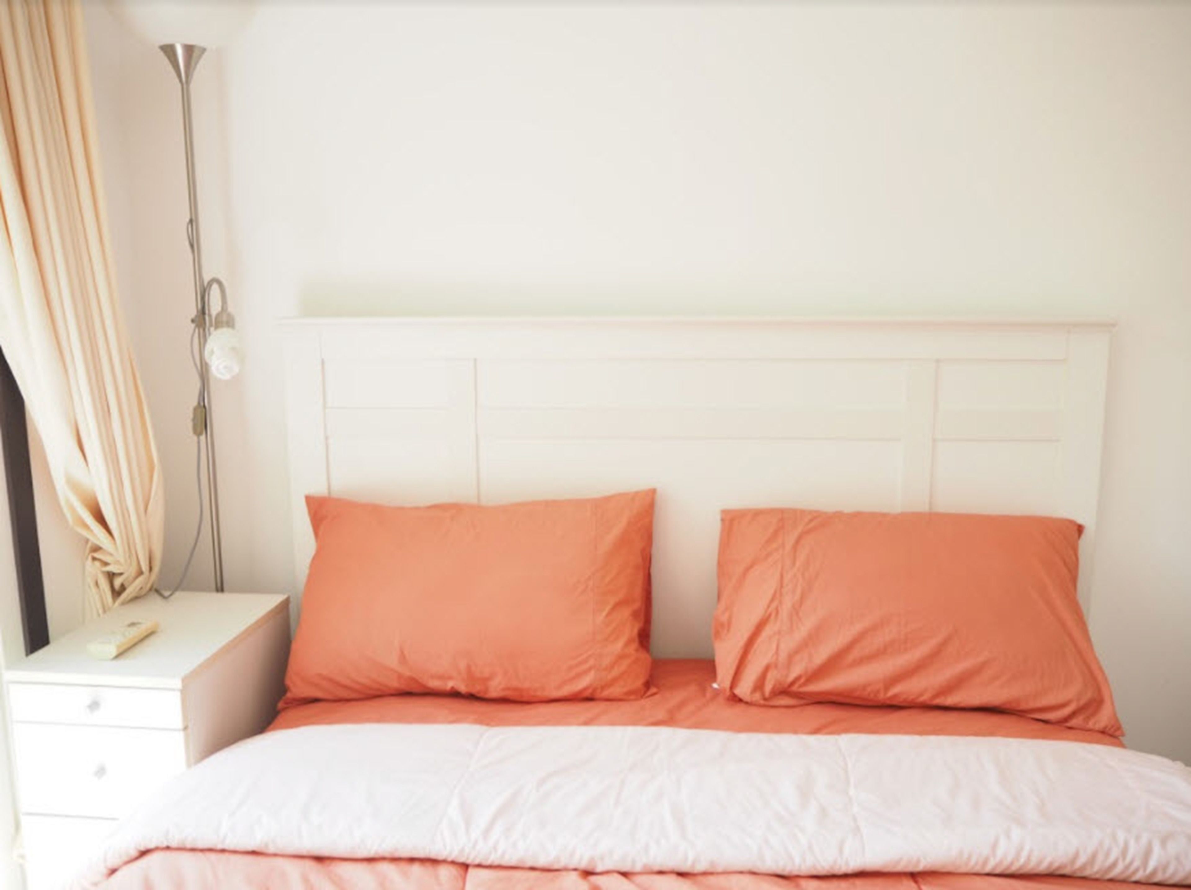 Een slaapkamer met een wit hoofdeinde, twee oranje kussens, een witte nachtkastje en een lamp.
