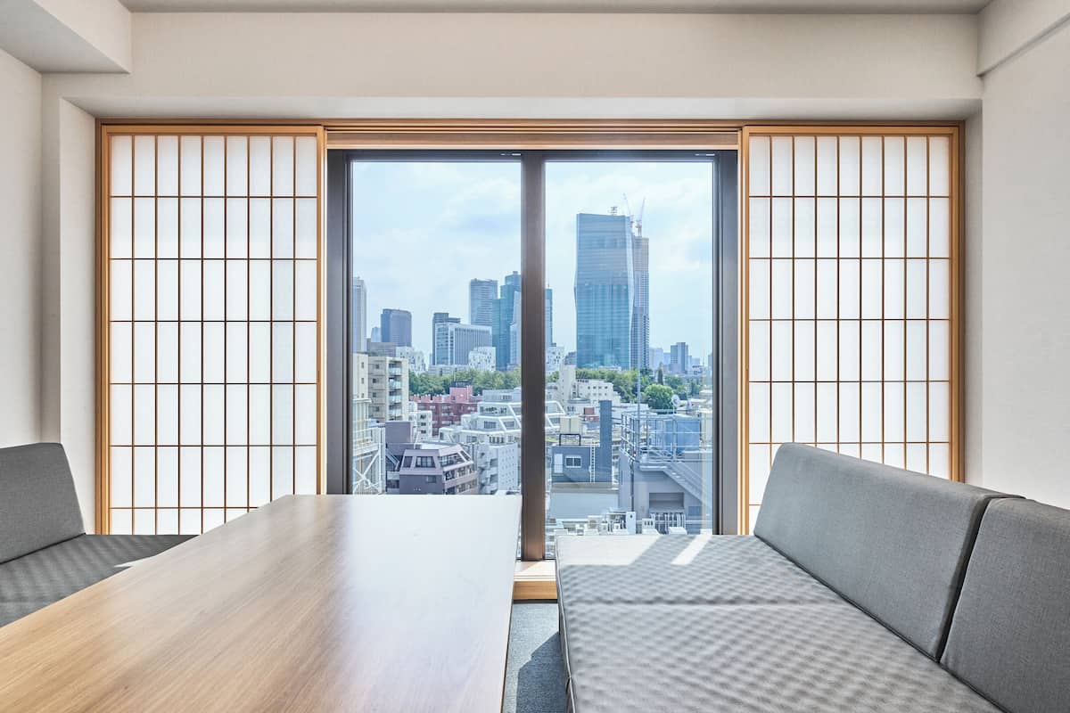 A modern hotel room with a large wooden table, two grey sofas, and a sliding glass door leading to a city view.
