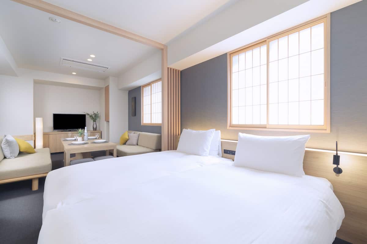 A hotel room with a large bed, a desk with a flat-screen TV, a sofa, and a window with blinds.