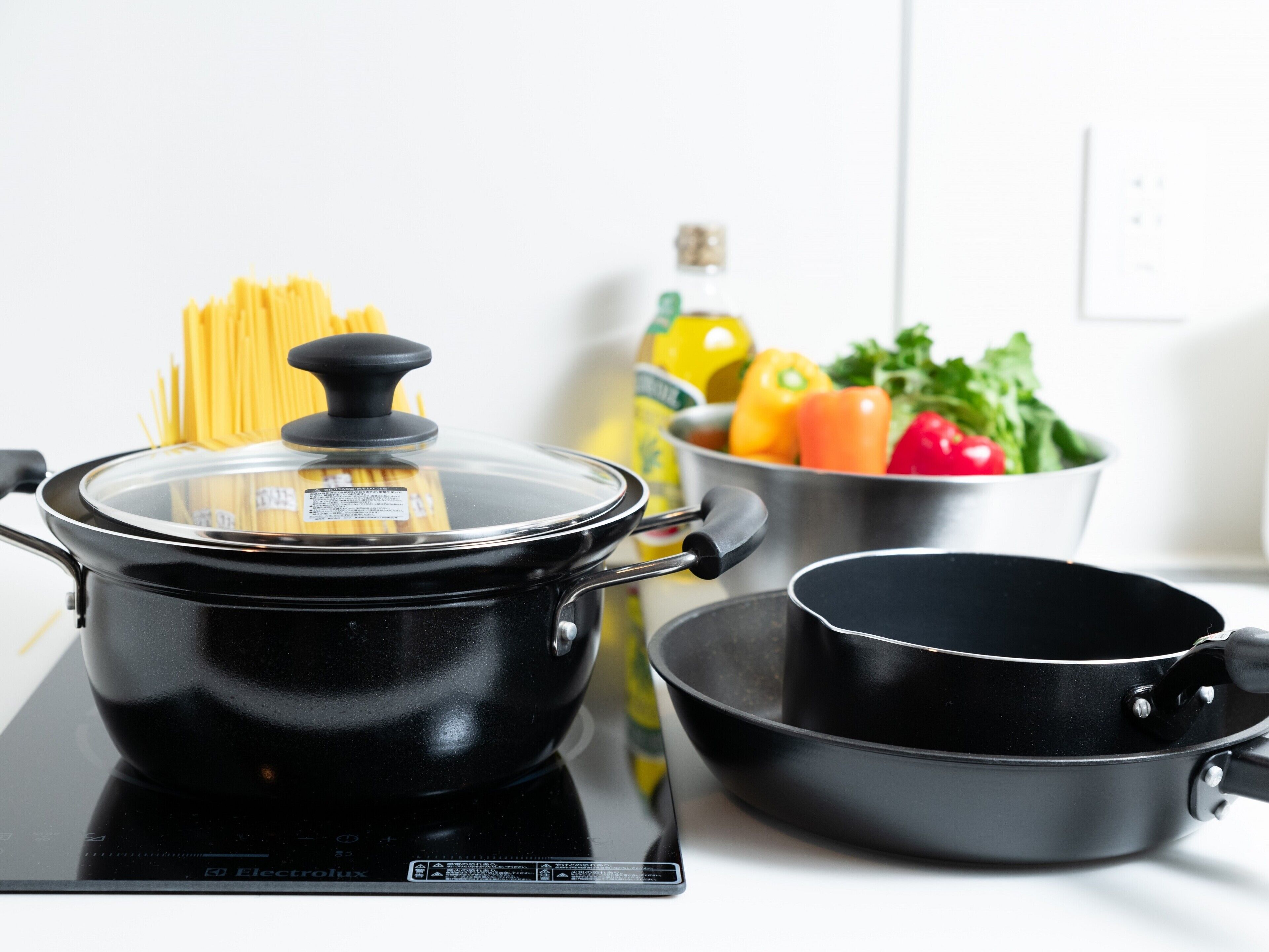 A kitchen with a black electric stove, a pot with a lid, a pan, and a bowl of vegetables.