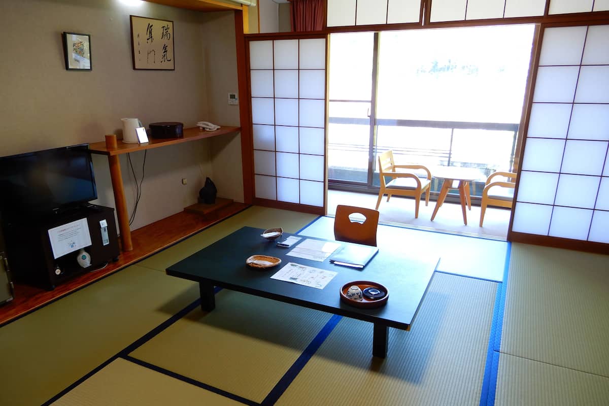 A traditional Japanese room with tatami flooring, a low table with documents, a television, and a view through shoji screens.