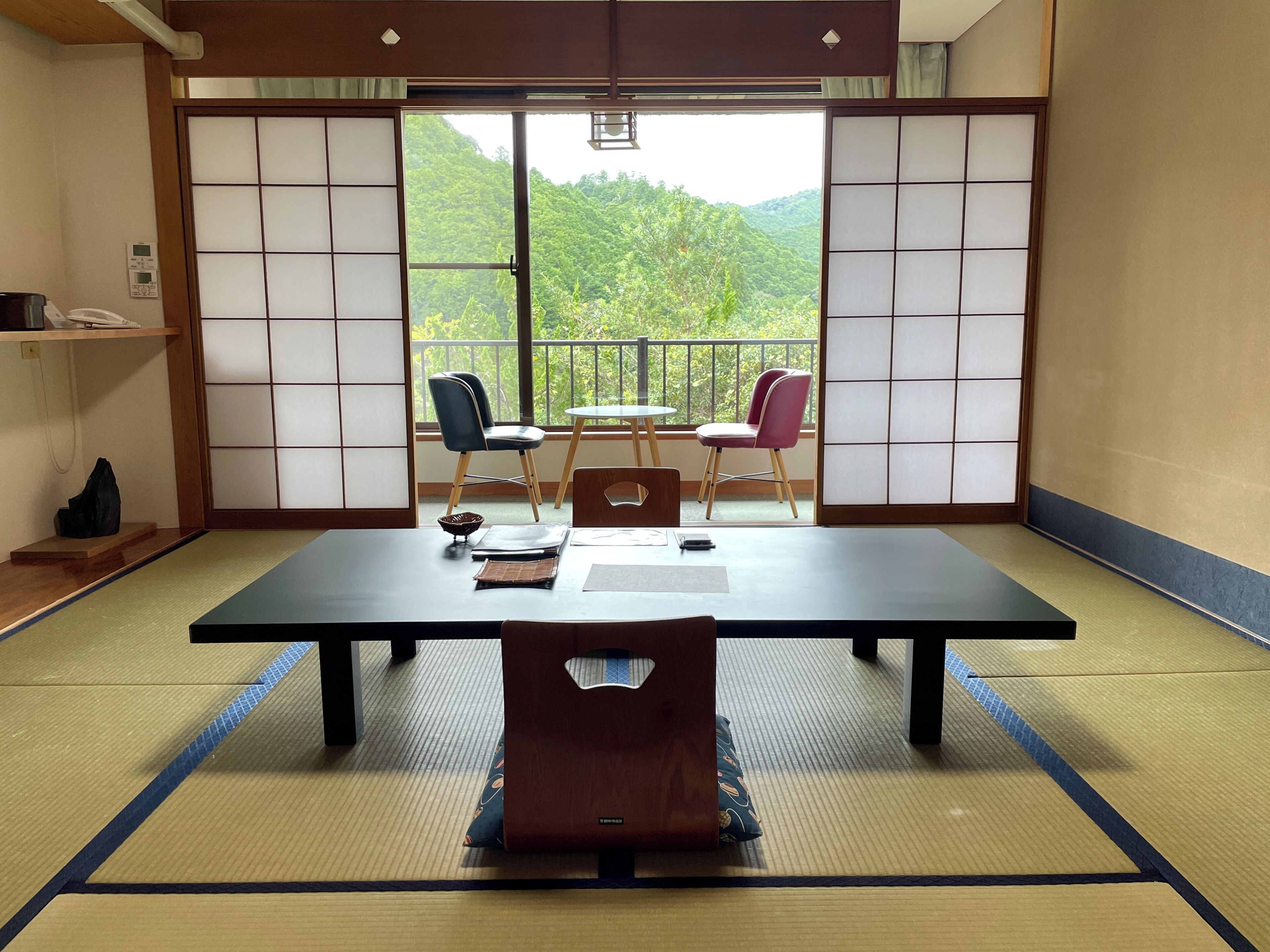 A tatami mat room with a low table, two chairs, a small table with a chair, and a view of greenery through shoji screens.