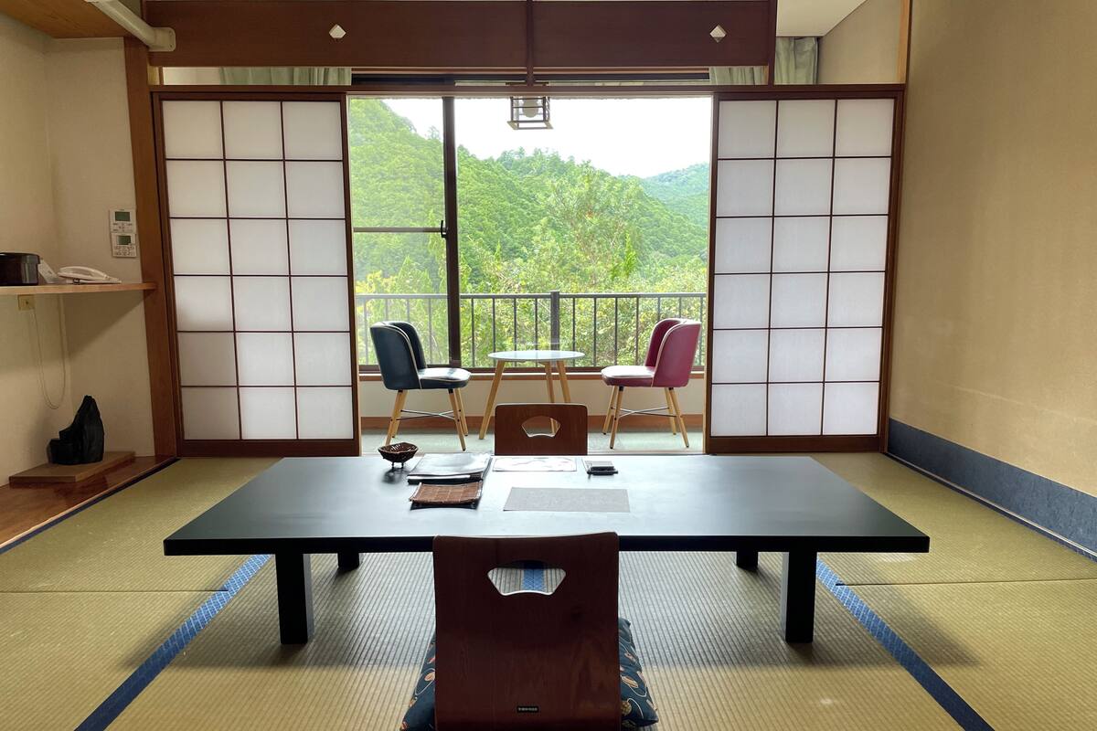 A tatami mat room with a low table, two chairs, a small table with a chair, and a view of greenery through shoji screens.