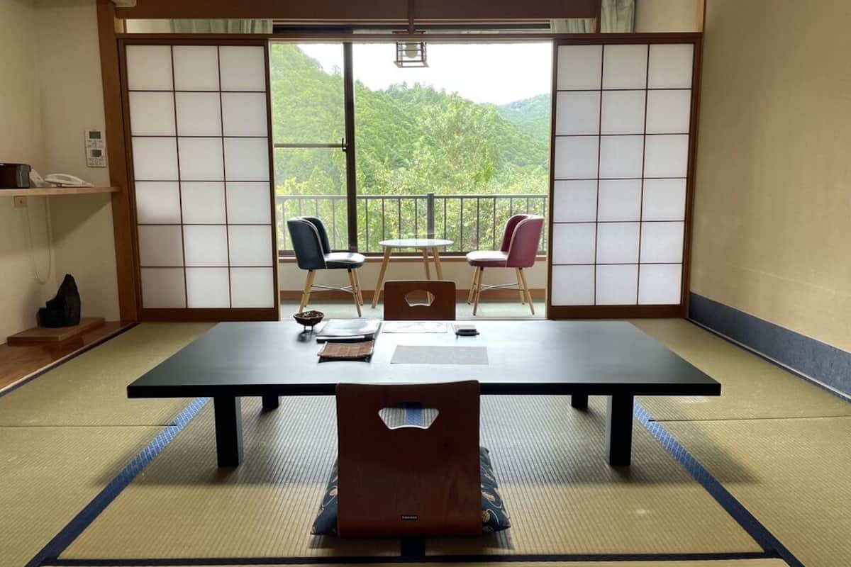 A tatami mat room with a low table, two chairs, a small table, and a view of greenery through a sliding door.