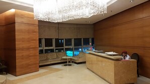 A modern reception area with a wooden reception desk, a seating area with sofas, and a large window with a view of the city at night. - Regalia Studio 109 Suites (Kuala Lumpur)