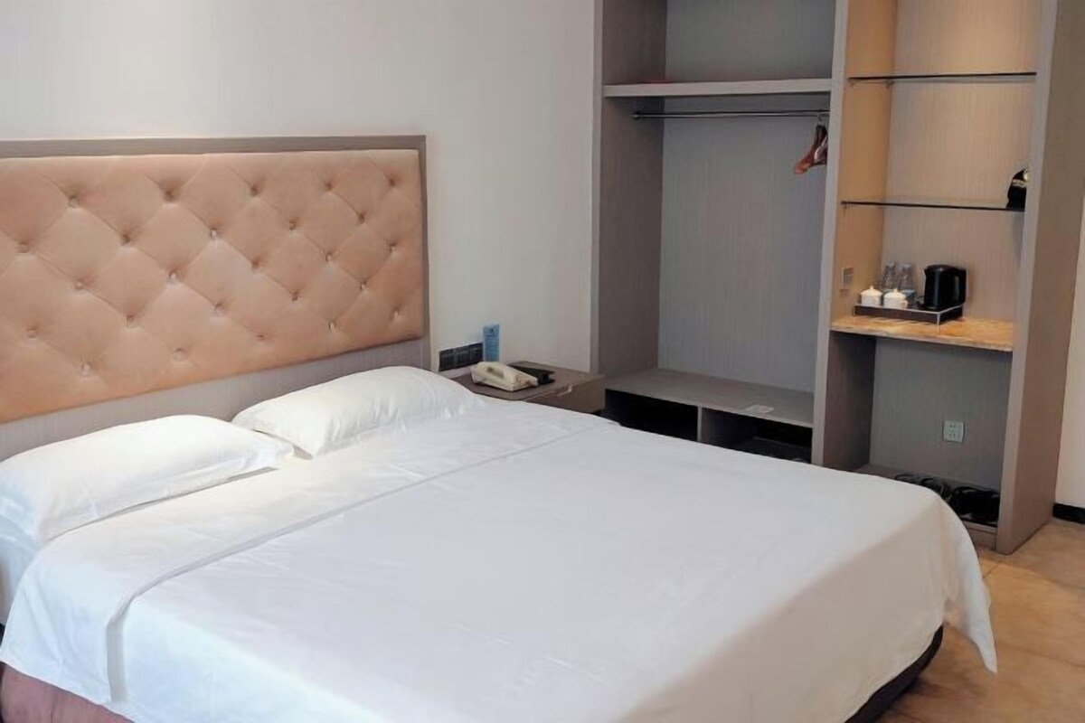 A hotel room with a large bed, a headboard, a bedside table, a wardrobe, and a shelf with items.
