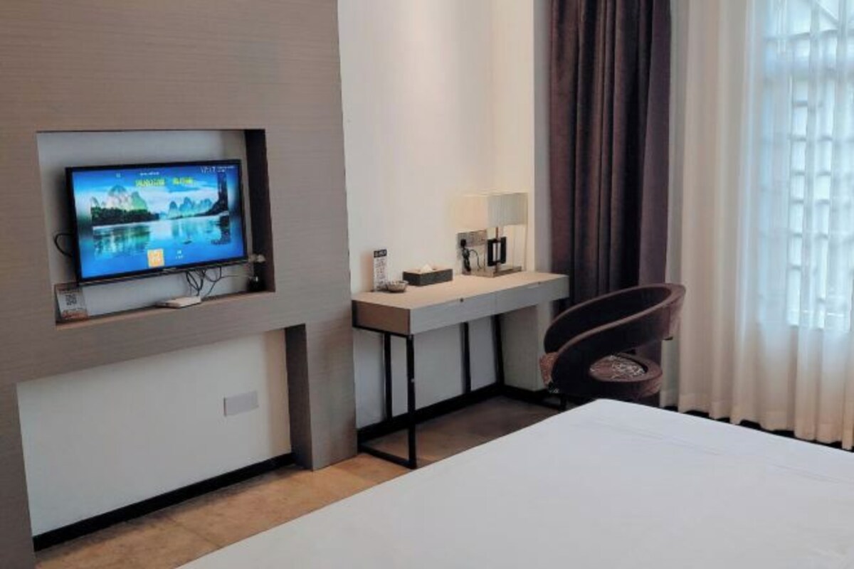 A hotel room with a bed, a desk with a computer, a chair, a TV mounted on the wall, and a window with curtains.