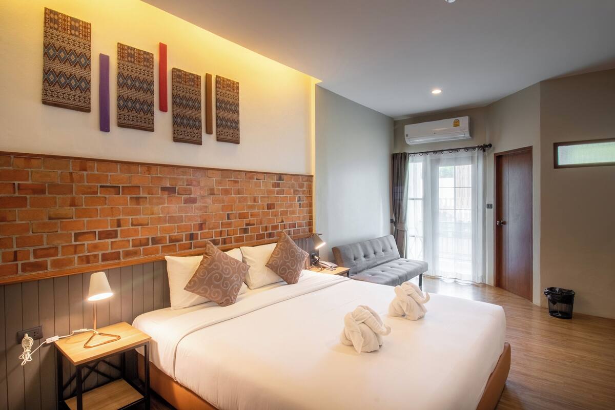A hotel room with a brick headboard, a bed with white linens, a grey sofa, and wall-mounted artwork.