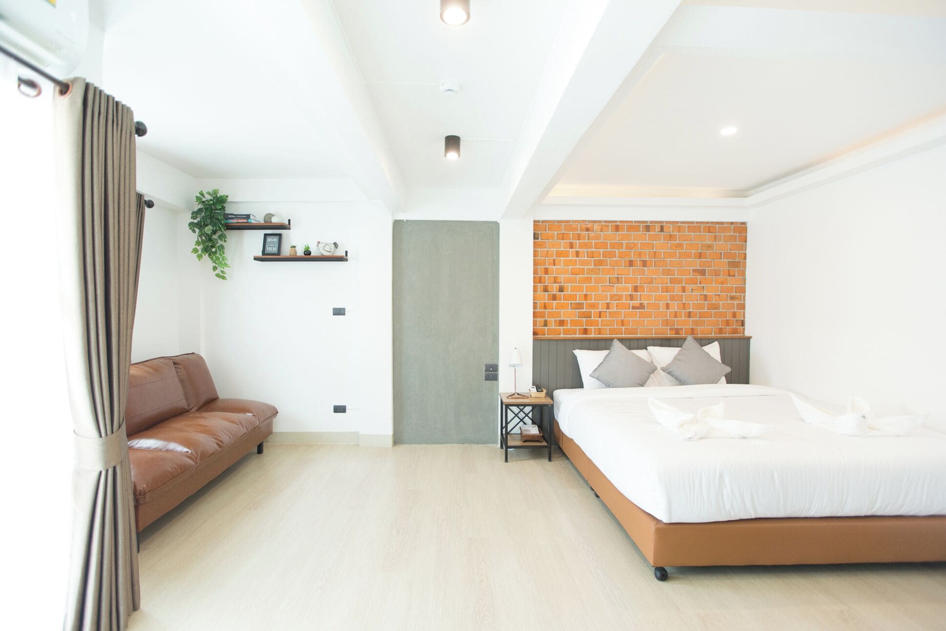 Un dormitorio moderno con una pared de ladrillo decorativa, un sofá de cuero marrón, una cama con ropa blanca y una mesita de noche con una lámpara.