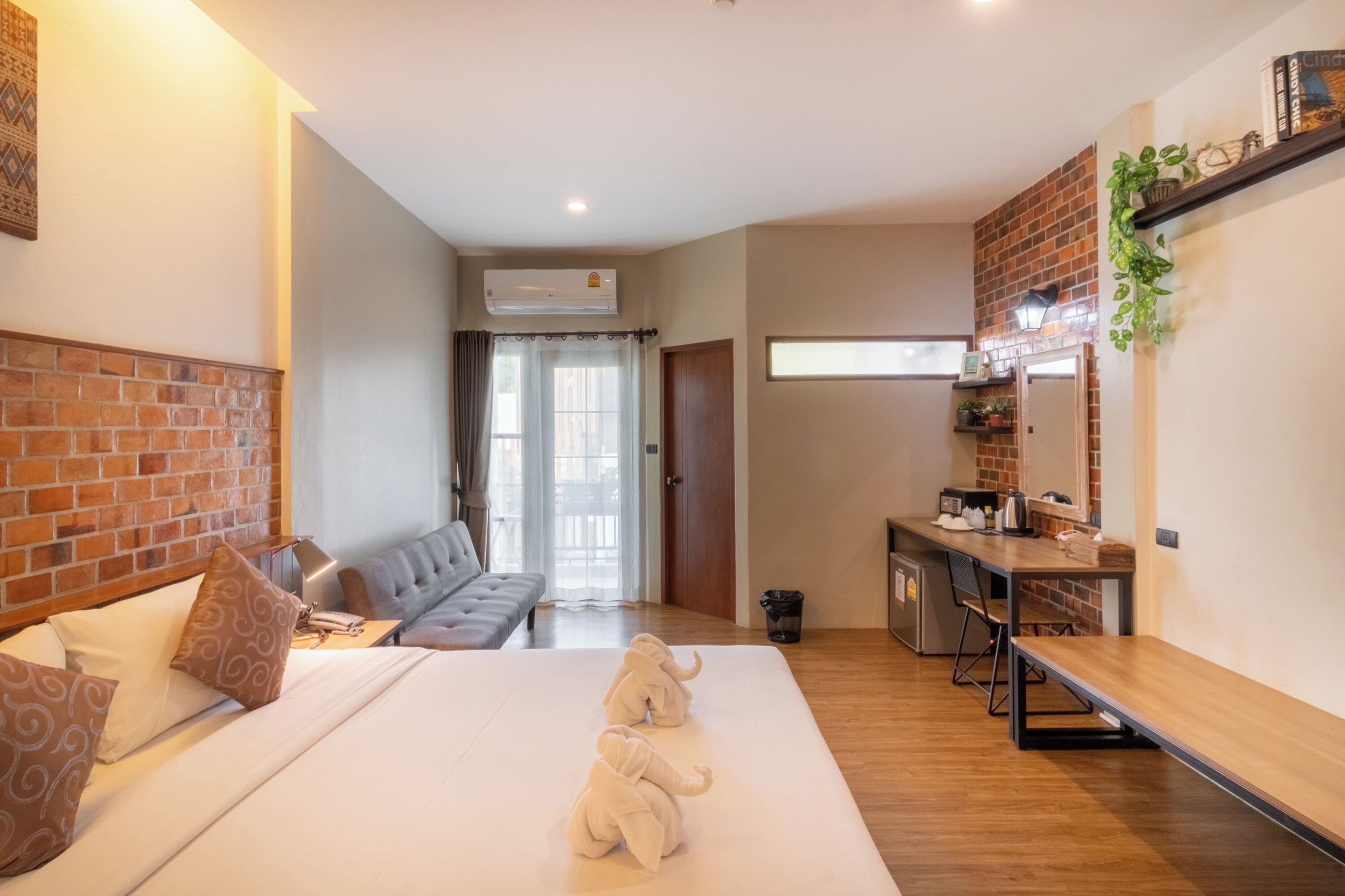 Una camera d'albergo con una parete decorata in mattoni, un letto con copripiumino bianco, un divano grigio, una piccola cucina e un'ampia finestra.