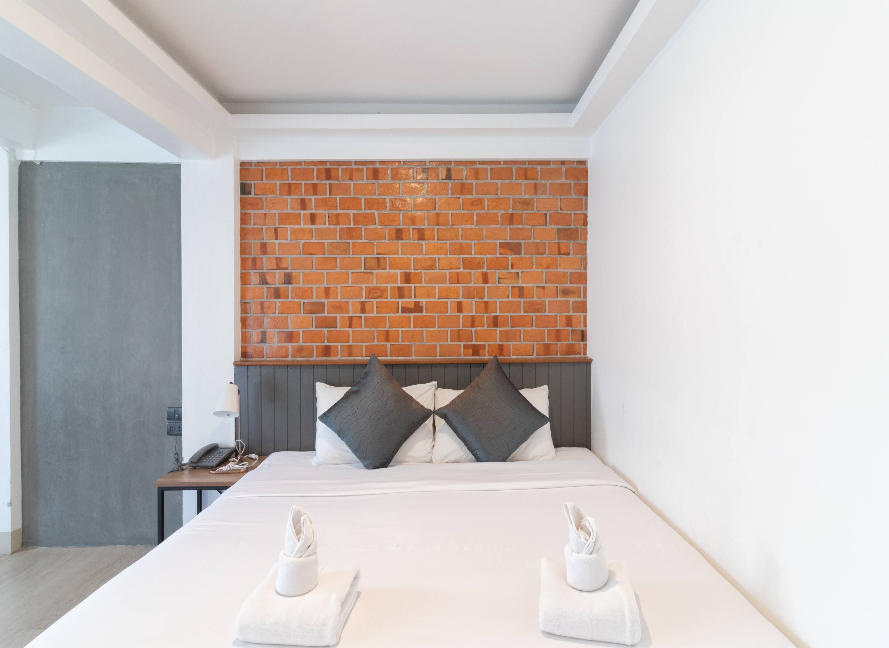 Een hotelkamer met een hoofdeinde van baksteenmotief, een opgemaakt bed met twee kussens, en een nachtkastje met een telefoon en een lamp.