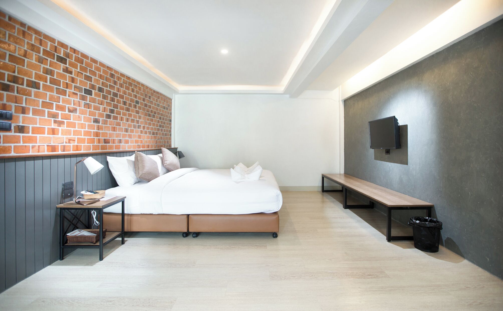 A modern bedroom with a brick wall, a bed with white bedding, a bench, a desk with a lamp, and a TV mounted on the wall.