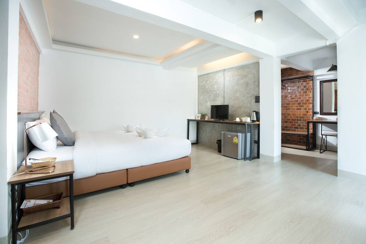A modern hotel room with a large bed, a desk with a television, a small kitchenette, and a brick wall.