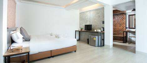 A modern hotel room with a large bed, a desk with a television, a small kitchenette, and a brick wall.