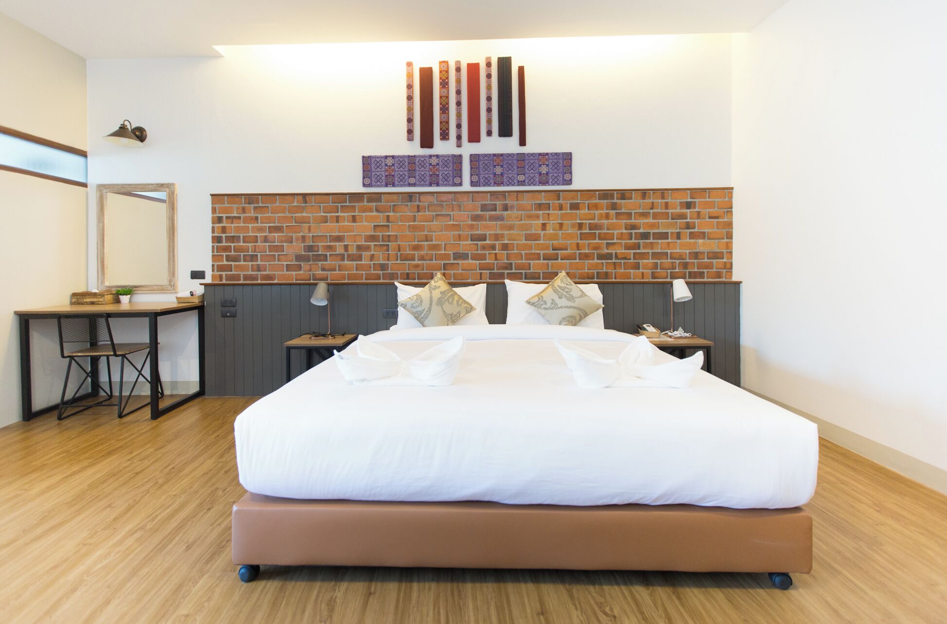 Une chambre d’hôtel moderne avec un mur d’accent en briques, un grand lit avec du linge de lit blanc, un bureau avec une chaise et un miroir.