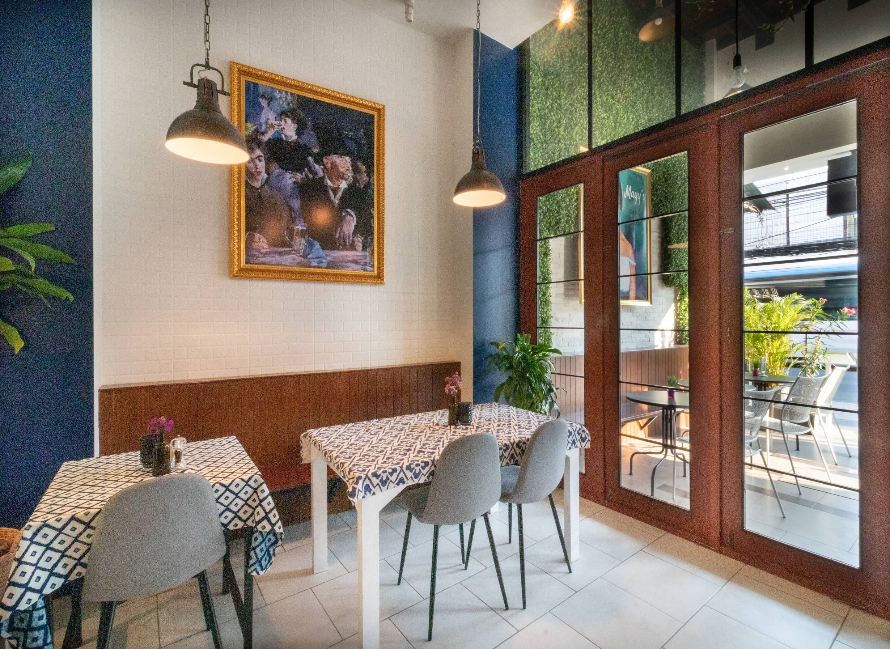 Interior de restaurante con zona de comedor que incluye mesas con manteles estampados y sillas, un cuadro grande en la pared y vistas al área de mesas al aire libre a través de puertas de vidrio.