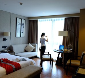 A hotel room with a bed, a sofa, a desk with a laptop, a chair, and a large window with curtains.