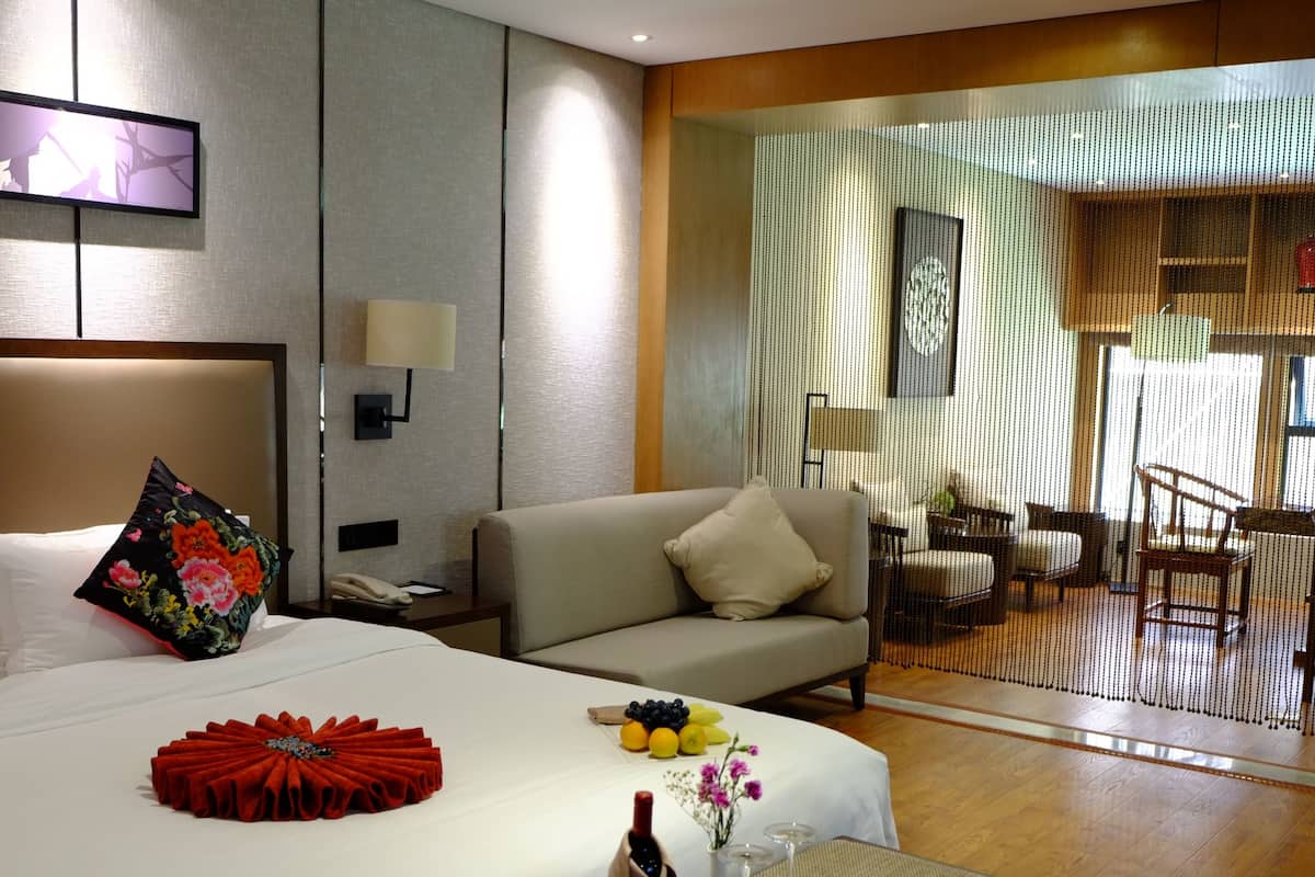 A hotel room with a bed, a bedside table, a nightstand, a sofa, a dining table with fruit and wine, and a view of the outdoor area.