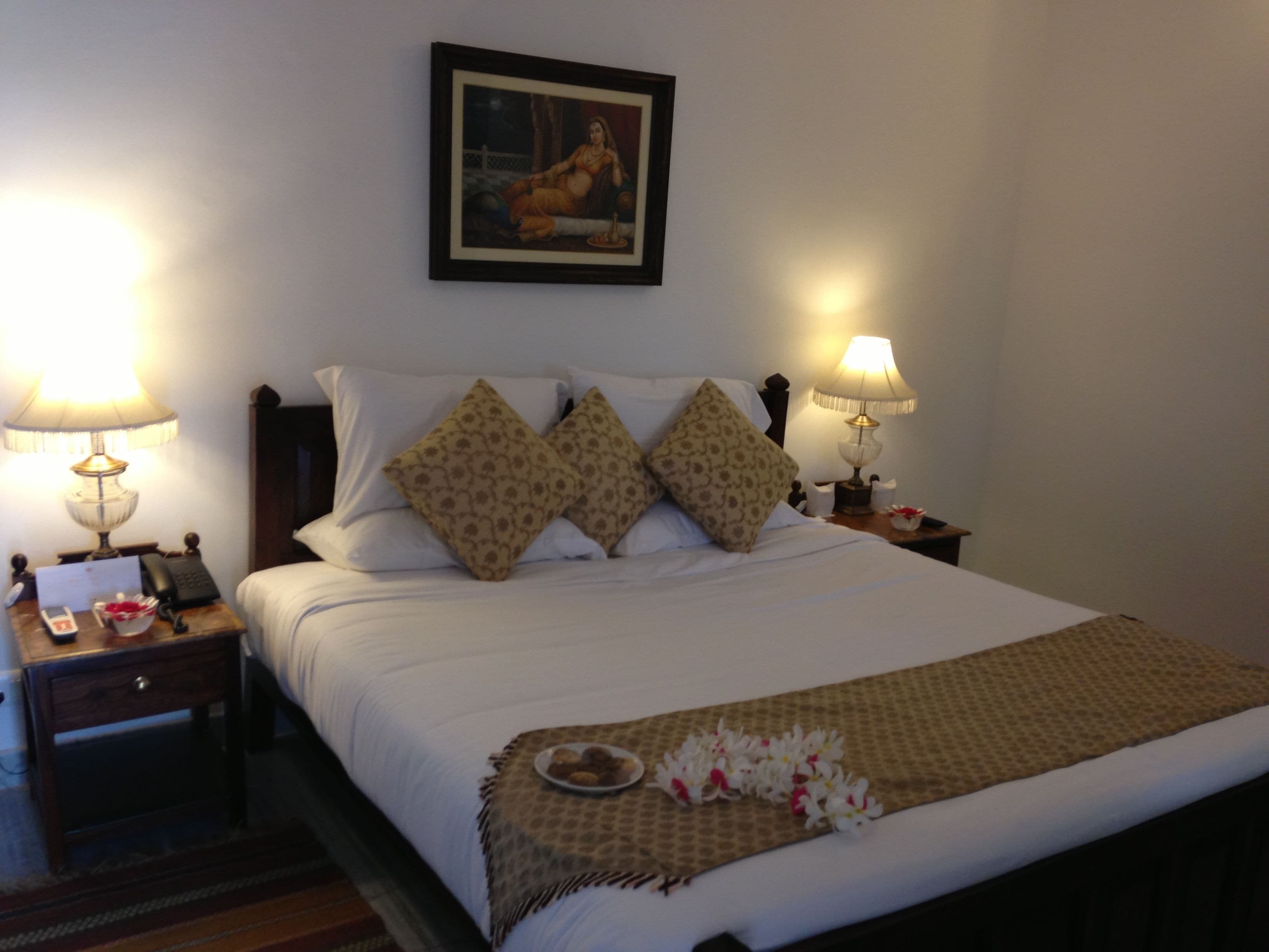 A neatly made bed with patterned pillows, two bedside lamps, a framed picture on the wall, and a small table with a phone and a bowl of flowers.