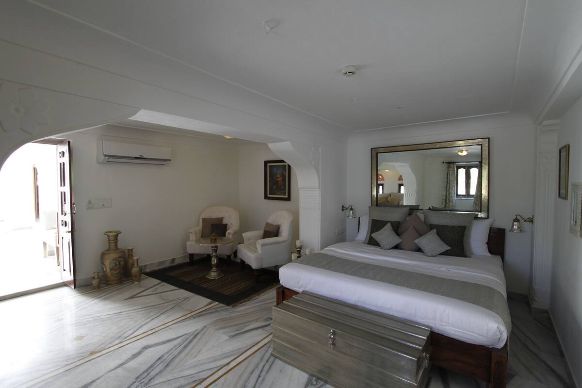 Une chambre spacieuse avec un grand lit, un coin salon avec deux fauteuils, un sol en marbre et un grand miroir.