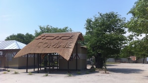 A thatched-roof structure with the word "GOMBE" written on it, surrounded by trees and a clear sky.