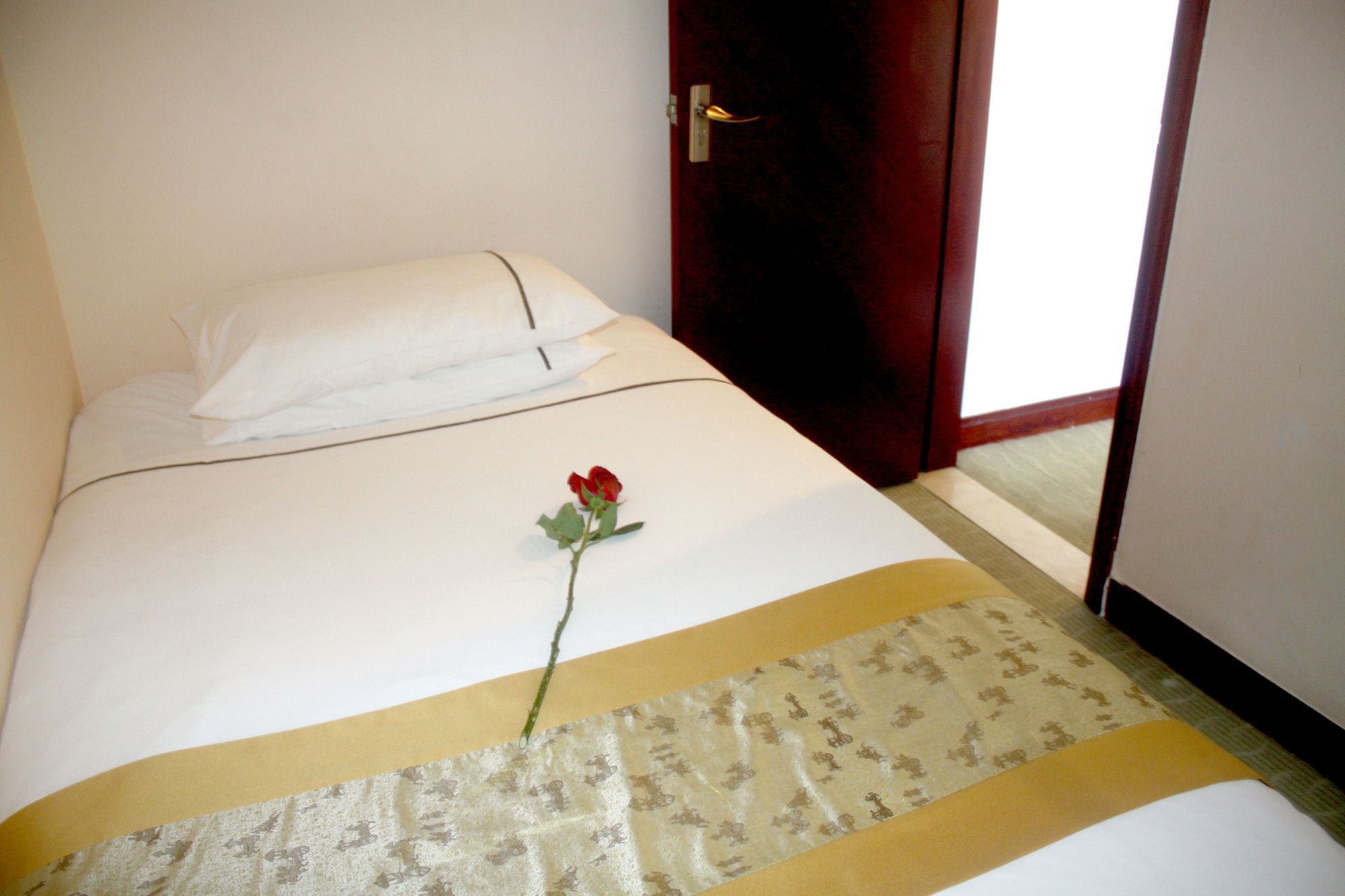 A neatly made bed with white linens, a single red rose, and a patterned runner.