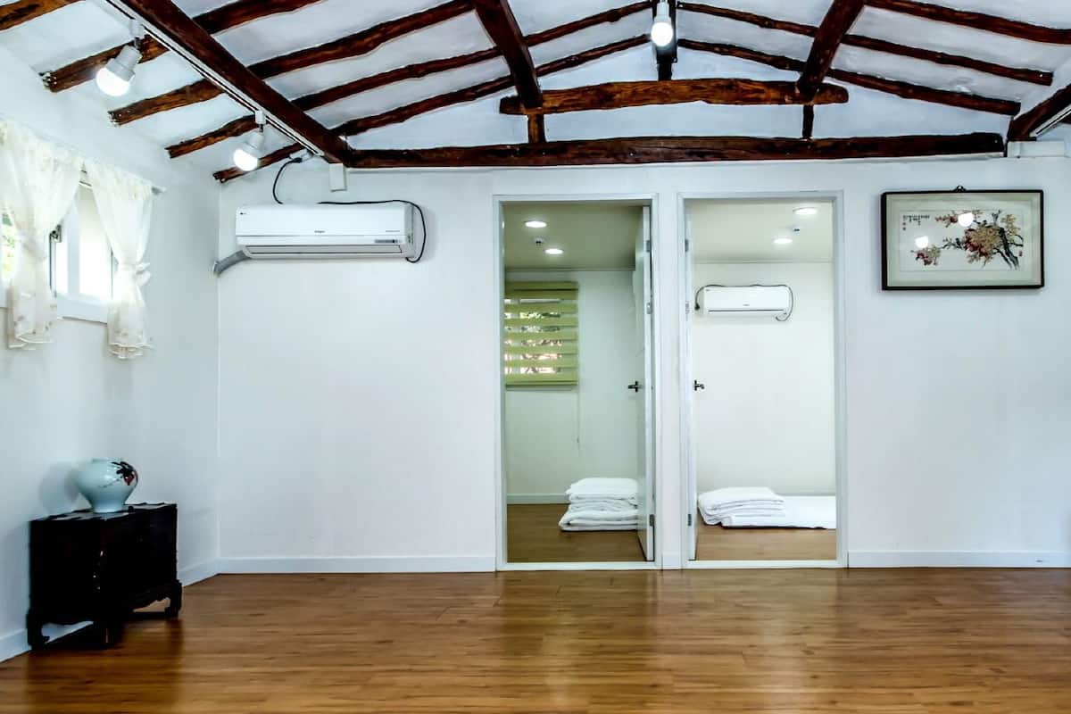 A room with wooden flooring, white walls, a ceiling with exposed beams, an air conditioning unit, a door leading to a bathroom, a framed picture, a coat rack, and a small table with a vase.