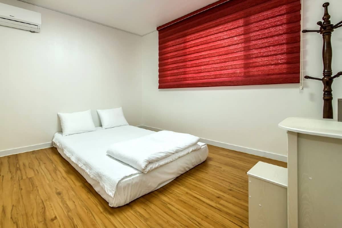 A bedroom with a wooden floor, a white bed with two pillows, a red window blind, and a white air conditioning unit on the wall.