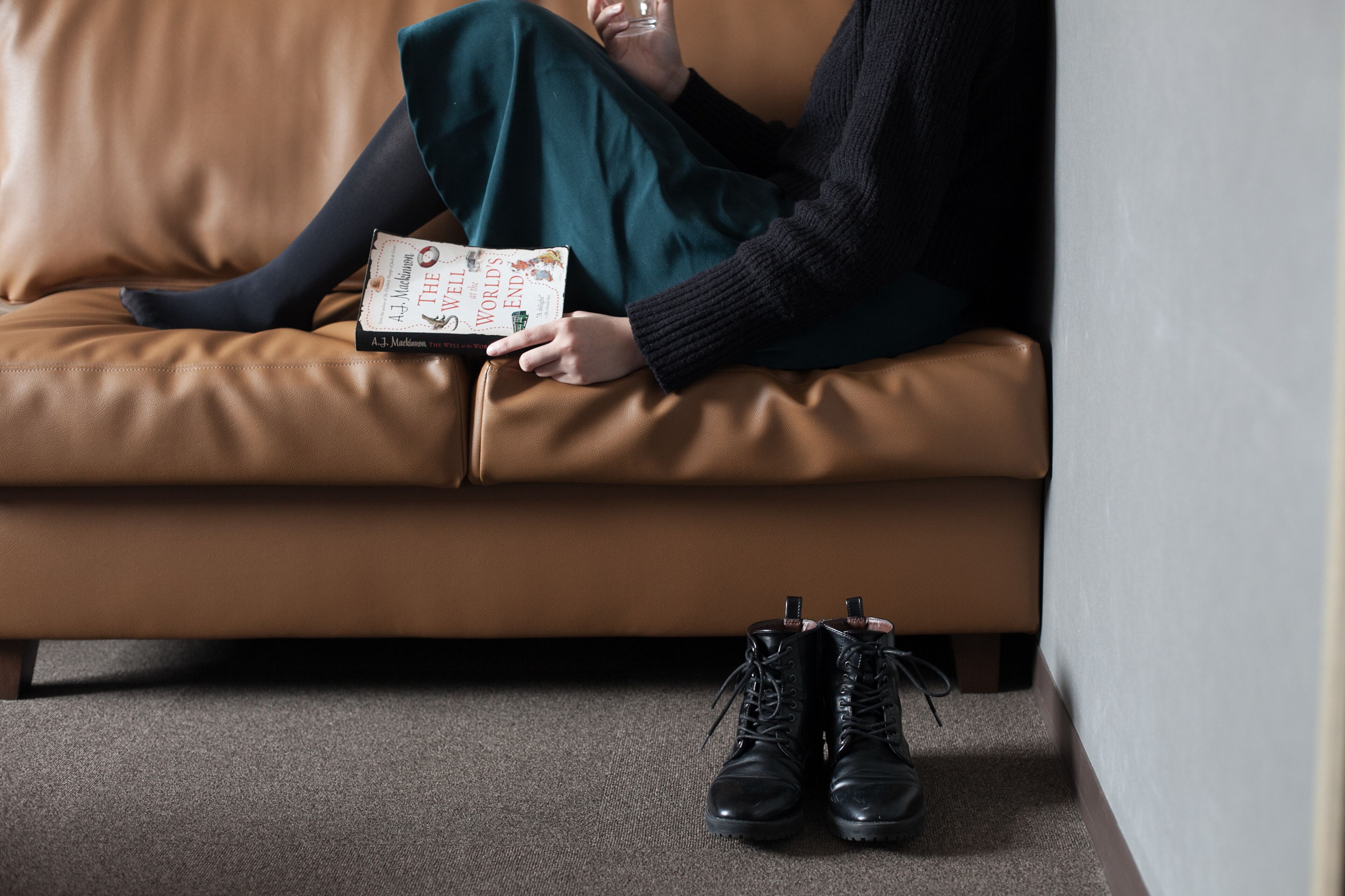Una persona sentada en un sofá de cuero marrón leyendo un libro, con unas botas negras a sus pies.