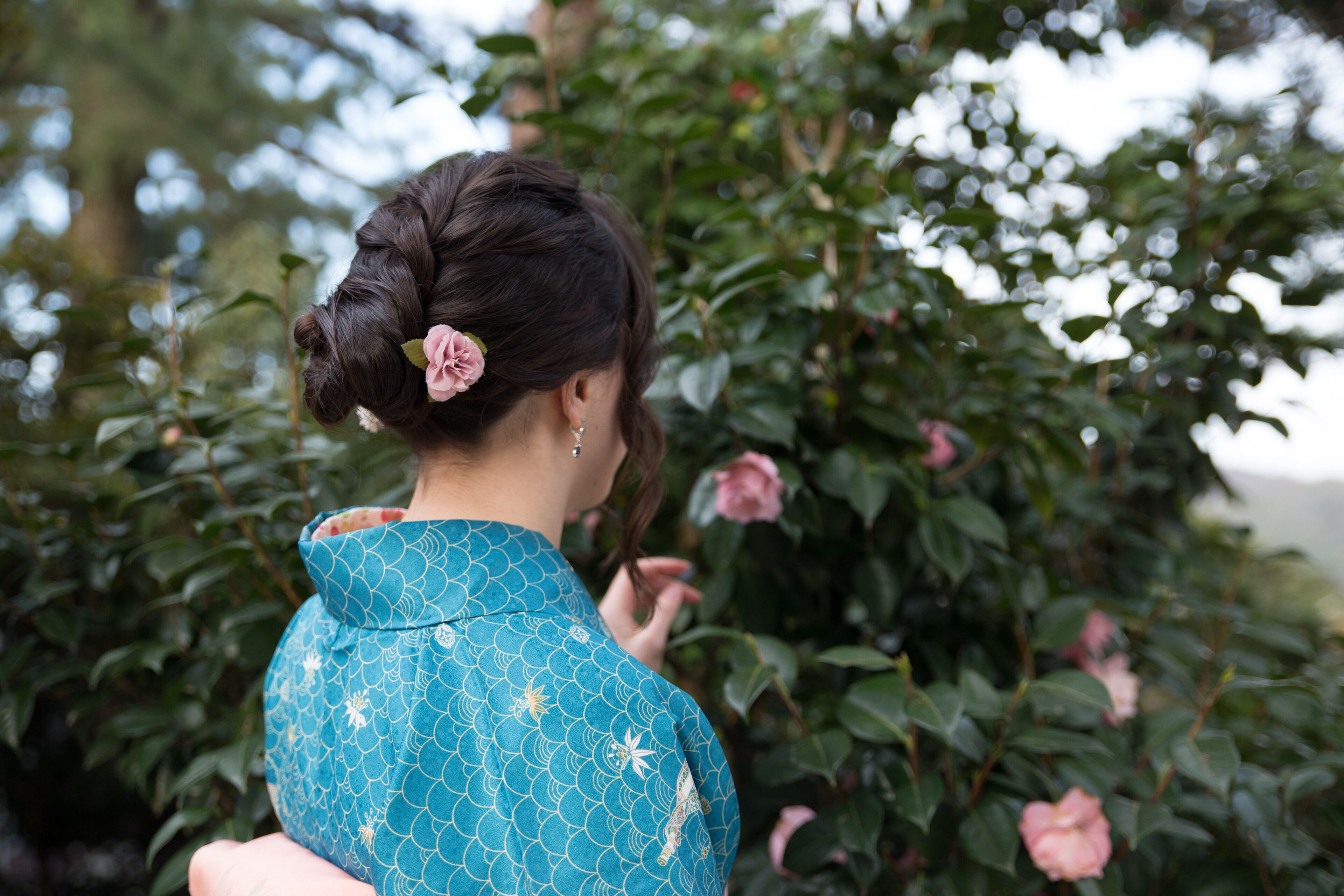 A person in a blue patterned garment standing in front of a bush with pink flowers.